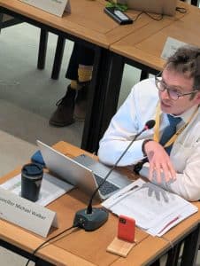 New Reform UK councillor, Michael Walker sat alone at a table. He has a laptop and a phone propped upright and some papers in front of him. He is gesturing with and open downward palm and he has his mouth open as he speaks.