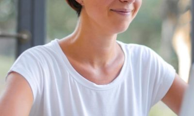 woman wears an opaque white t-shirt for women; she is typing on a laptop and sitting in front of a window with trees and grass behind her.