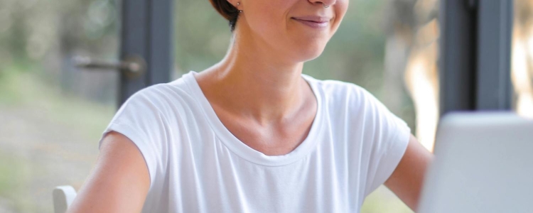 woman wears an opaque white t-shirt for women; she is typing on a laptop and sitting in front of a window with trees and grass behind her.
