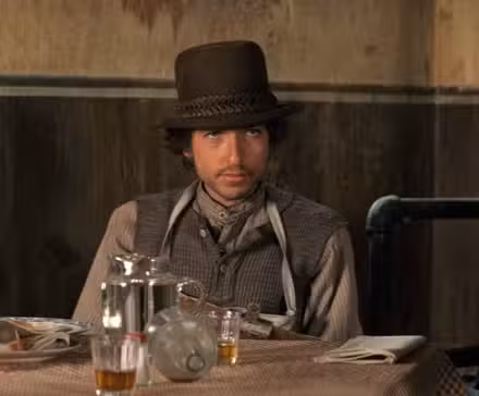 Alias (Bob Dylan) sits at a bar table surrounded by liquor bottles and glasses in 'Pat Garrett and Billy the Kid'