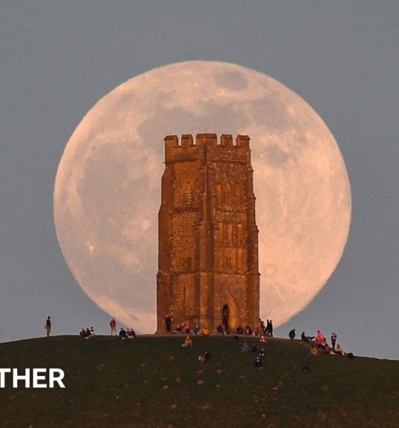 A huge pink hued Moon sits in a grey sky, behind the tower on Glastonbury Tor with people milling around at the base