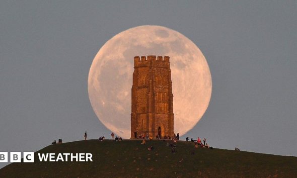 A huge pink hued Moon sits in a grey sky, behind the tower on Glastonbury Tor with people milling around at the base