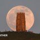 A huge pink hued Moon sits in a grey sky, behind the tower on Glastonbury Tor with people milling around at the base