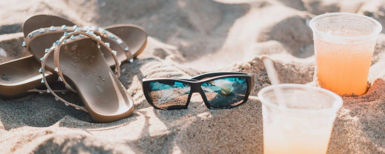 stock photo of a sandy beach with two plastic cups containing an orange drink, sunglasses, and a pair of sandals with studded straps