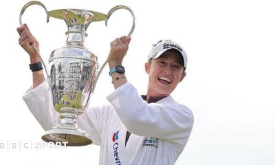 Nelly Korda is wearing a white dressing gown and baseball cap and holding the Chevron Championship in the air
