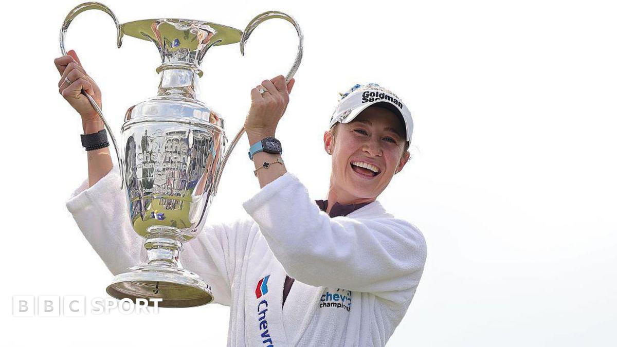 Nelly Korda is wearing a white dressing gown and baseball cap and holding the Chevron Championship in the air