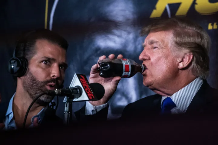 President Donald Trump drinks a soda as he hosts the Holyfield vs Belford boxing match live in 2021, next to his son Donald Trump Jr.