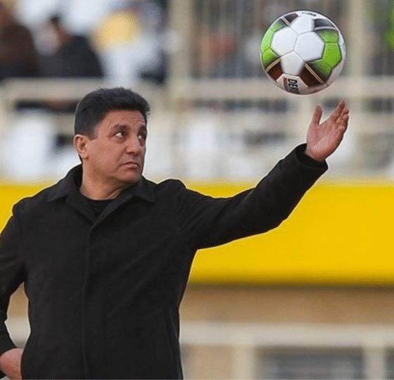 Iran national football team coach, Amir Ghalenoei, throws a football in the air with one hand while the other rests confidently in his pocket. He resembles a strong statue and stares at the ball in mid-air.
