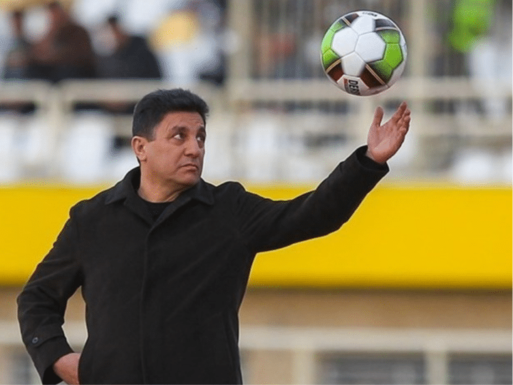 Iran national football team coach, Amir Ghalenoei, throws a football in the air with one hand while the other rests confidently in his pocket. He resembles a strong statue and stares at the ball in mid-air.