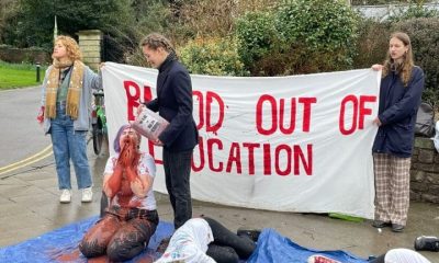 A Bristol University Arms Industry Blood out of Education protest in 2024. A student kneels on the ground covered in red liquid and another lies motionless. ‘Blood out of education’ is written in red on a banner behind them.