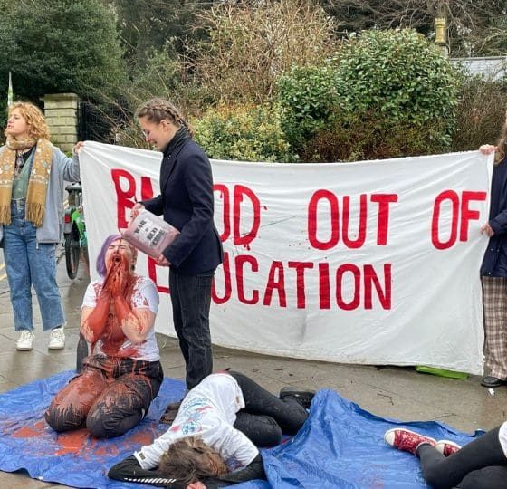 A Bristol University Arms Industry Blood out of Education protest in 2024. A student kneels on the ground covered in red liquid and another lies motionless. ‘Blood out of education’ is written in red on a banner behind them.