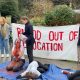A Bristol University Arms Industry Blood out of Education protest in 2024. A student kneels on the ground covered in red liquid and another lies motionless. ‘Blood out of education’ is written in red on a banner behind them.