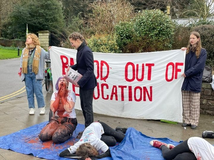 A Bristol University Arms Industry Blood out of Education protest in 2024. A student kneels on the ground covered in red liquid and another lies motionless. ‘Blood out of education’ is written in red on a banner behind them.