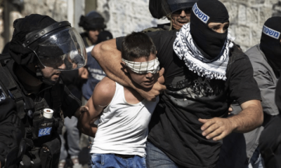 A child in Palestine, a boy, is gripped around his neck by an IOF member while blindfolded. His hands are held behind his back