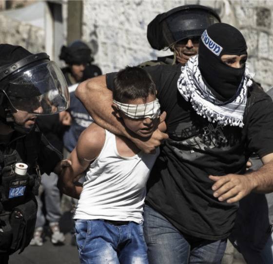 A child in Palestine, a boy, is gripped around his neck by an IOF member while blindfolded. His hands are held behind his back