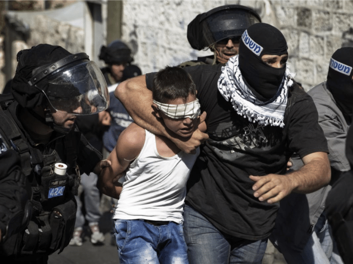 A child in Palestine, a boy, is gripped around his neck by an IOF member while blindfolded. His hands are held behind his back