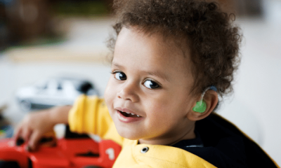 A deaf young boy with curly hair turns to smile at the camera with one hand on his plastic toy car. His ear shows his colourful hearing aid.