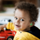 A deaf young boy with curly hair turns to smile at the camera with one hand on his plastic toy car. His ear shows his colourful hearing aid.
