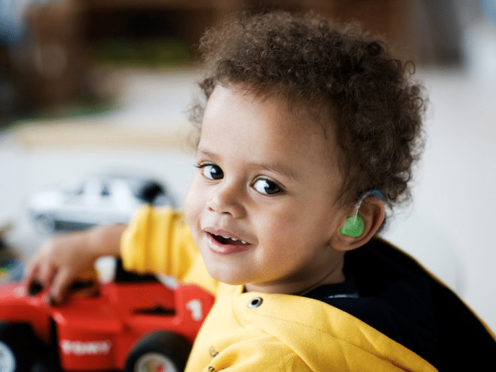 A deaf young boy with curly hair turns to smile at the camera with one hand on his plastic toy car. His ear shows his colourful hearing aid.