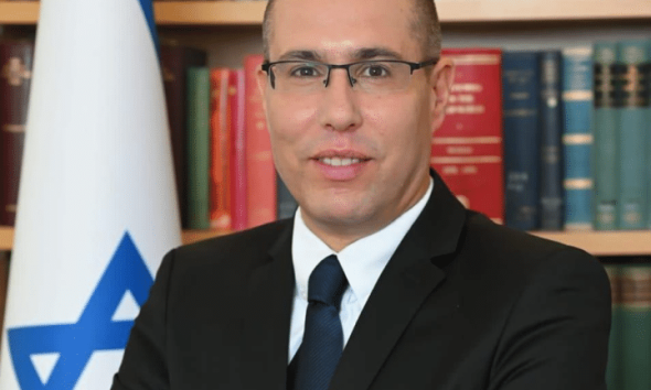 Eli Hazan, Benjamin Netanyahu's comms director smiles in a corporate headshot with the Israel flag and a packed bookshelf behind him