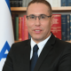 Eli Hazan, Benjamin Netanyahu's comms director smiles in a corporate headshot with the Israel flag and a packed bookshelf behind him