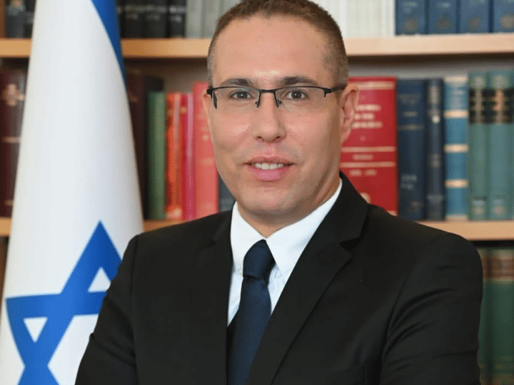 Eli Hazan, Benjamin Netanyahu's comms director smiles in a corporate headshot with the Israel flag and a packed bookshelf behind him