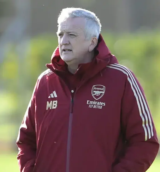 Mark Bonnick stands on the pitch wearing an Arsenal jacket with his hands in his pocket as though watching players in the distance