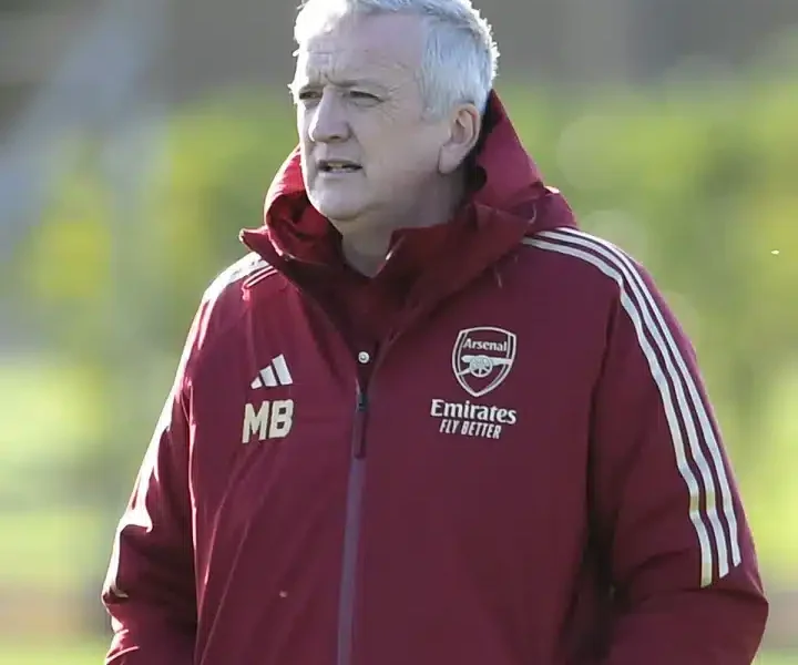 Mark Bonnick stands on the pitch wearing an Arsenal jacket with his hands in his pocket as though watching players in the distance
