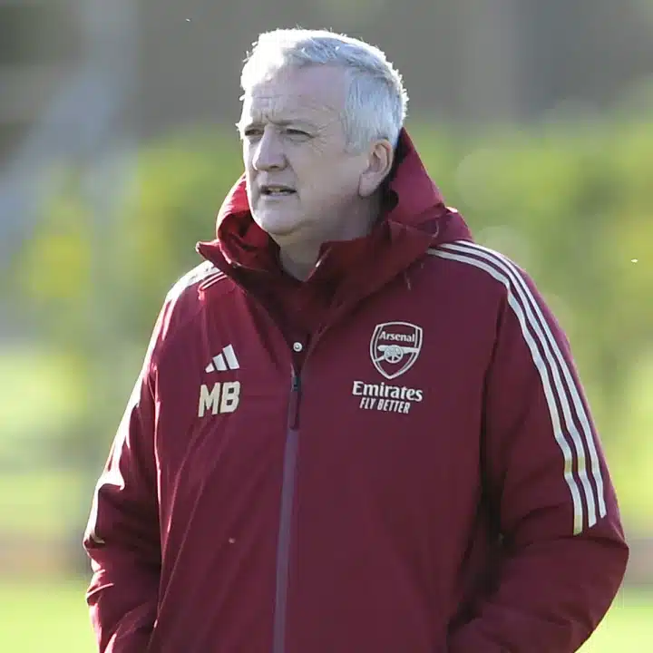 Mark Bonnick stands on the pitch wearing an Arsenal jacket with his hands in his pocket as though watching players in the distance