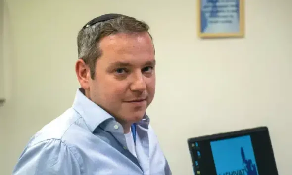 Shivat Zion founder and CEO, Shraga Evers, smiles with a laptop in the background displaying the charity's logo on screen