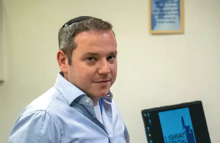 Shivat Zion founder and CEO, Shraga Evers, smiles with a laptop in the background displaying the charity's logo on screen