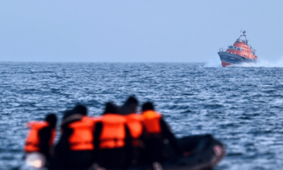A group of people wearing life jackets sit on an inflatable boat in the middle of the sea. A larger vessel in the distance sails towards them