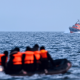 A group of people wearing life jackets sit on an inflatable boat in the middle of the sea. A larger vessel in the distance sails towards them