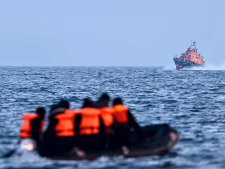 A group of people wearing life jackets sit on an inflatable boat in the middle of the sea. A larger vessel in the distance sails towards them