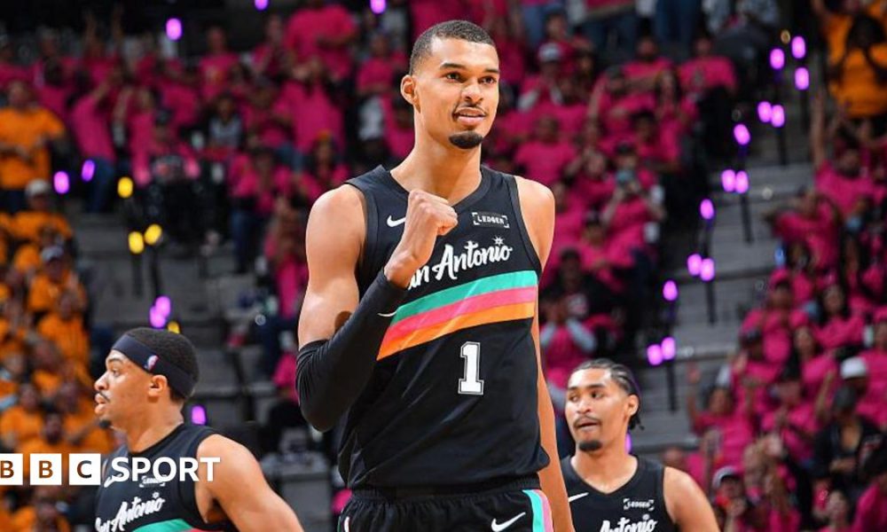 Victor Wembanyama, wearing a black San Antonio vest with a turquoise, pink, and orange stripe across the chest, punches his right fist in celebration and smiles