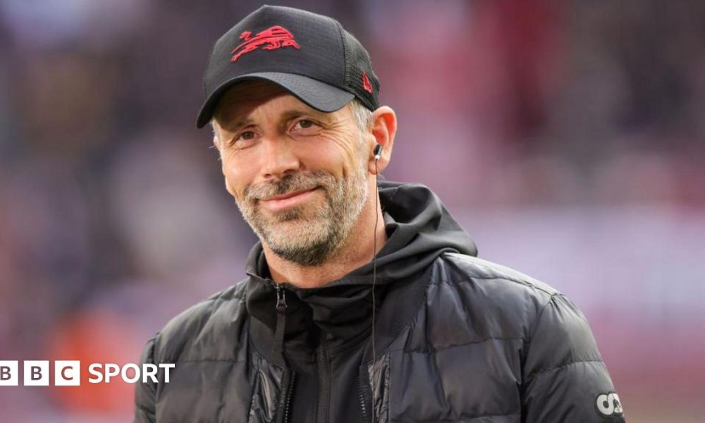 Marco Rose smiles and is wearing a baseball cap and a black coat