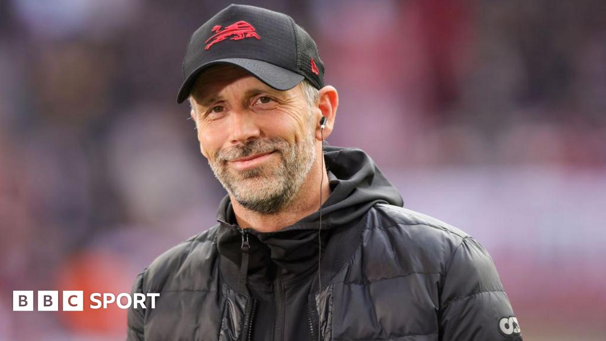 Marco Rose smiles and is wearing a baseball cap and a black coat