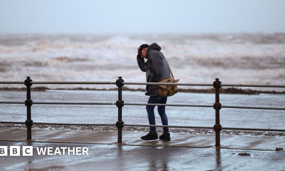 A man clutches his hat while walking along a windy seafront with rough waves in the background