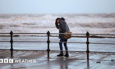 A man clutches his hat while walking along a windy seafront with rough waves in the background