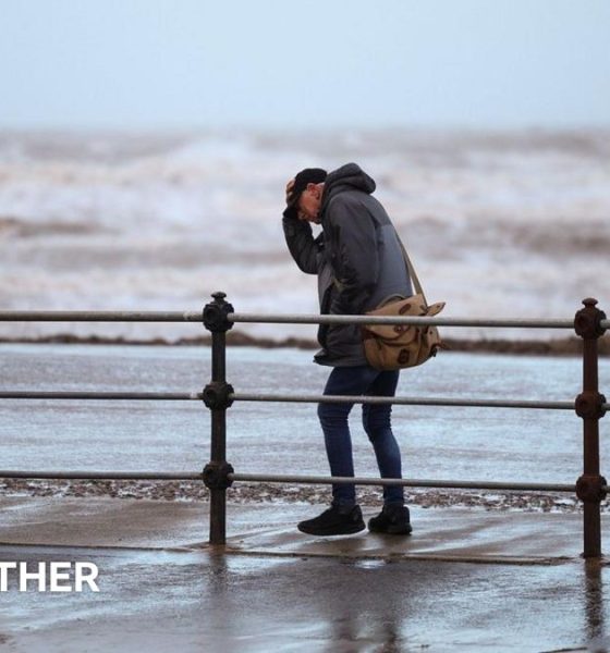 A man clutches his hat while walking along a windy seafront with rough waves in the background