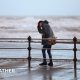 A man clutches his hat while walking along a windy seafront with rough waves in the background