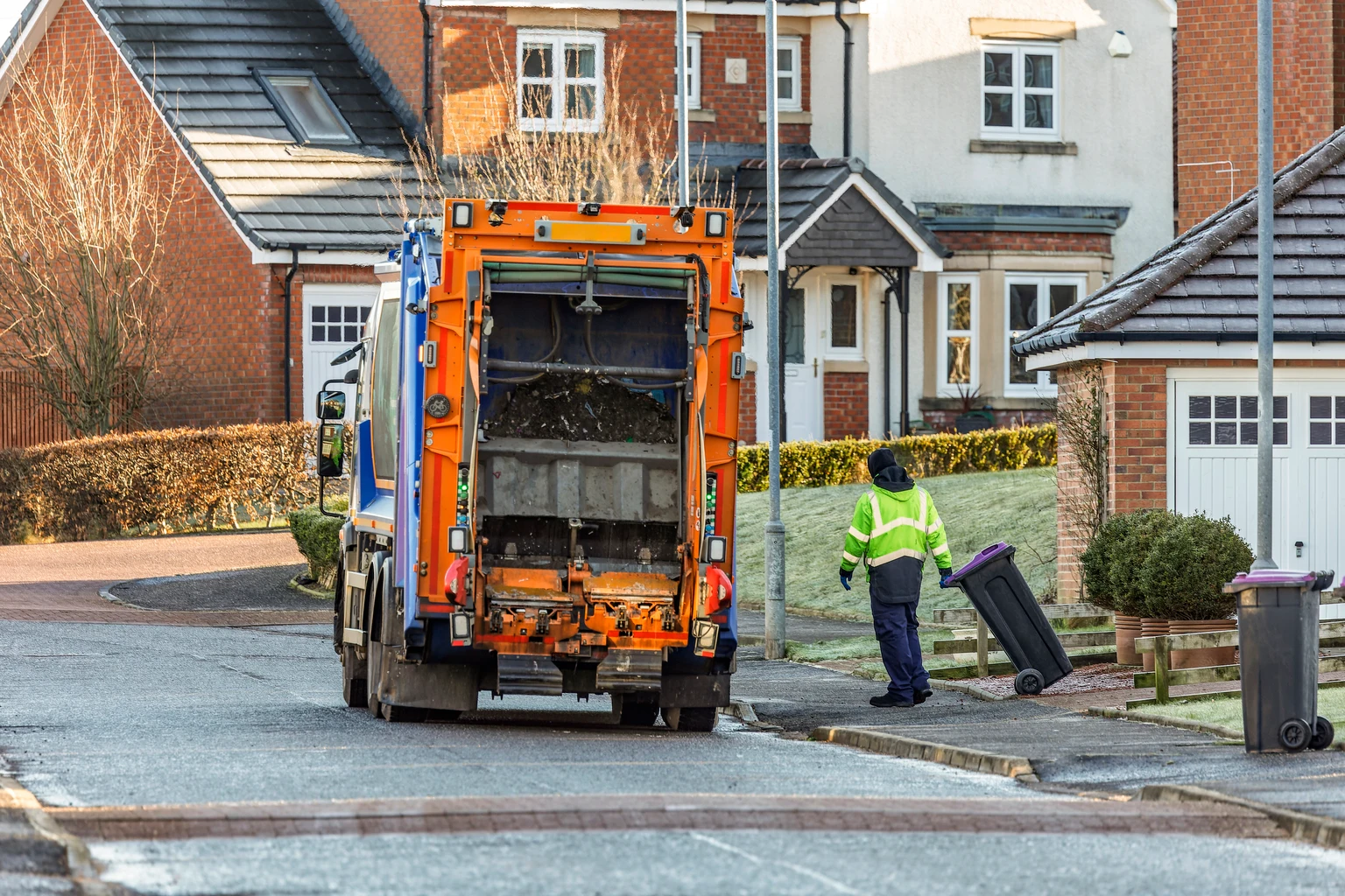 Municipal Refuse Truck and Worker in Hi-Vis Vest Collecting Residential Waste Recycling Wheelie Bins in Suburban Residential Street