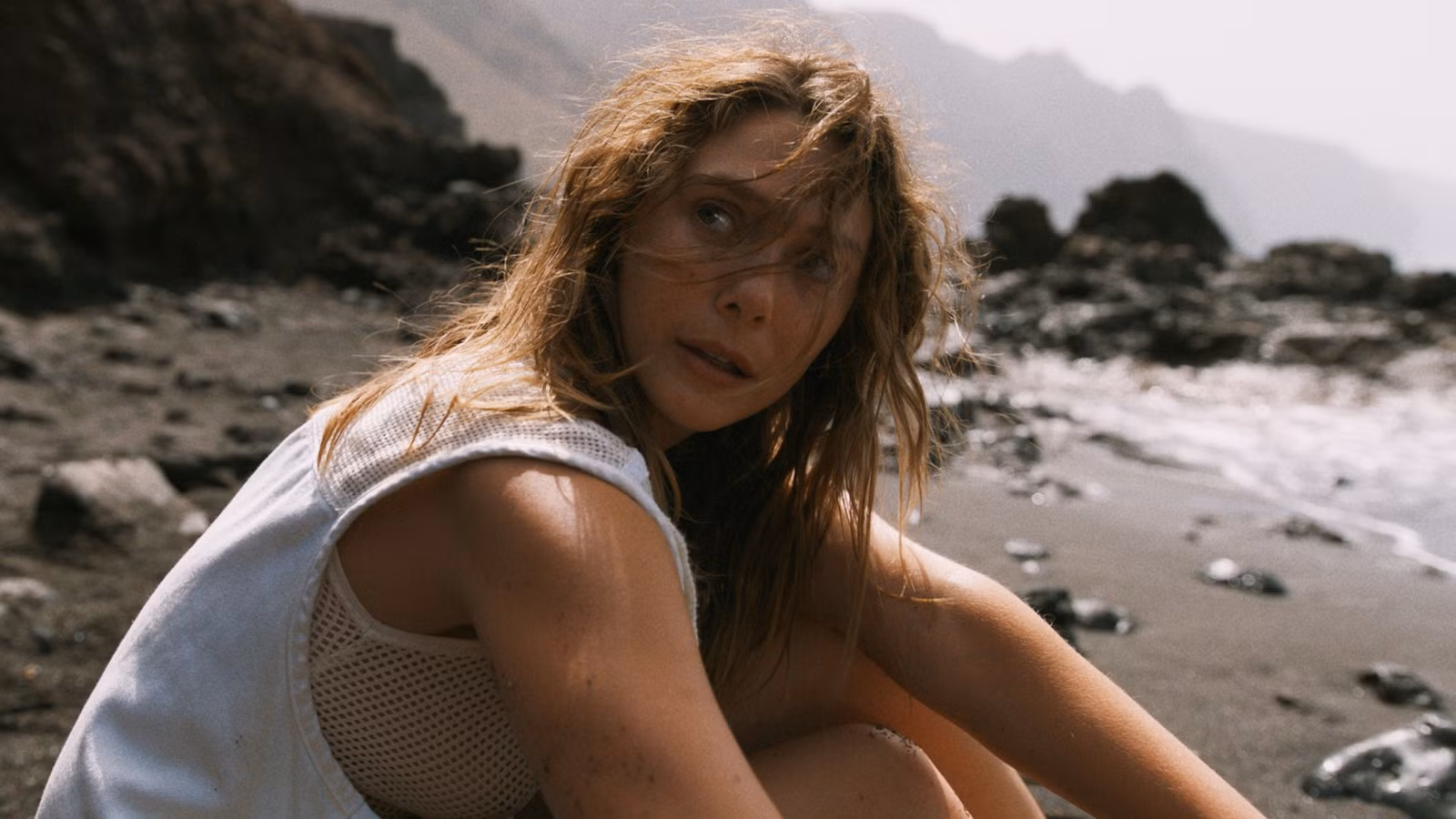 Elizbeth Olsen as Mia sitting in the sand on the beach and looking over her right shoulder in The Assessment