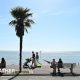 sunny scene of a coastal path where people are sitting on benches with other people walking along next to the glistening sea with blue skies overhead