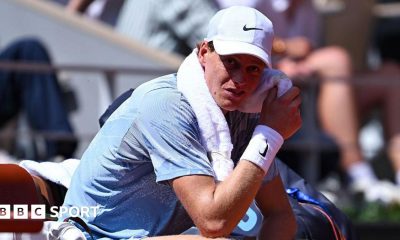 Jannik Sinner, wearing a grey t-shirt and white cap, sits in his chair with a towel around his neck wearing a pained expression