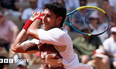 Adolfo Daniel Vallejo, wearing a white patterned t-shirt, plays a backhand with his yellow, blue and black racquet held in two hands over his right shoulder