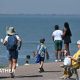 A beach prominade with one lady sitting on a bench while others are walking and looking out to sea