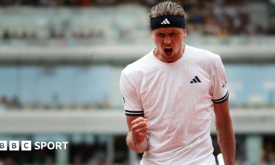 Alexander Zverev, wearing a white t-shirt and black headband, roars with his mouth wide open and clenches his right fist in celebration