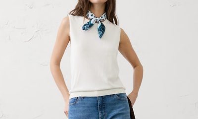 A woman wearing a white tank, white-and-blue bandana around her neck, and blue jeans. She is carrying a black jacket.