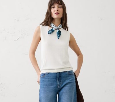 A woman wearing a white tank, white-and-blue bandana around her neck, and blue jeans. She is carrying a black jacket.